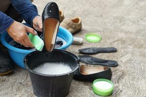 Closeup hands use brush to clean and wash black shoes in black bucket and blue bowl outdoor. Concept, take care, maintenance footwears from dirt and bad smell for using long time. Hygiene and sanitary photo
