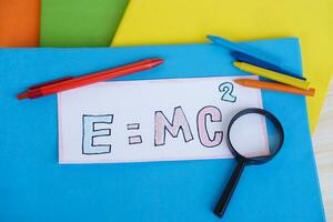 Paper card of formula E MC2 on colorful books with pen, crayons and magnifying glass. Concept, Einstein formula. Mass Energy Equivalence. Education, physics subject. photo