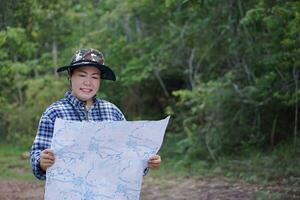 asiático mujer explorador sostiene papel mapa a encuesta ruta en bosque. concepto, de viaje, naturaleza exploración. ecología estudiar. pasatiempo actividad, aventuras estilo de vida. explorar ambiente. foto