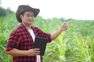 Handsome Asian man farmer is in garden, wears hat, red plaid shirt, holds  smart tablet. Feel confident, proud in crops. Concept, Agriculture occupation, inspect, survey and research. Copy space. photo