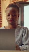 Vertical video Portrait of teleworker in warm lit room working on online report tasks at desk, solving duties remotely at home. African american woman with blogger job, e business. Handheld shot.