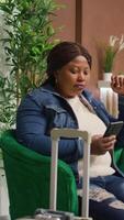 Vertical video While waiting for check in time, tourist texting messages on mobile phone in luxurious lounge area. African american woman preparing to register into room, having holiday reservation.