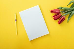 A blank sheet in a notebook with a pen and red tulips with green leaves lie on a yellow background. photo