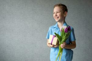 un linda chico sonrisas y soportes con un regalo y tulipanes dar un ramo de flores de flores foto