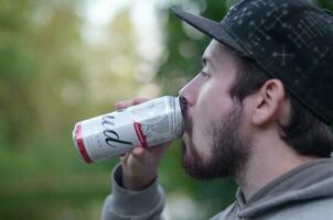 Young bearded man drinks Budweiser Bud beer in aluminium can on green trees blurred background photo