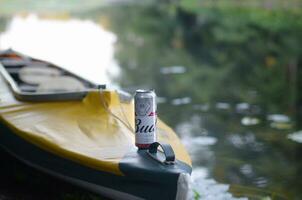 Budweiser Bud beer can on yellow kayak outdoors in the river and green trees blurred background photo