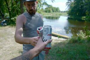 Young man raise Budweiser Bud beer can with male friend on blurred river with kayak and trees photo