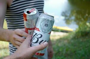 Young man raise Budweiser Bud beer can with male friend on blurred river with kayak and trees photo