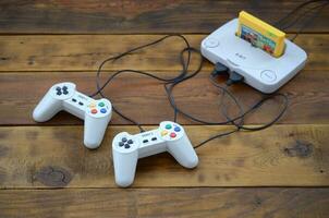 Old 8-bit video game console with game cartridge and two gaming joysticks on wooden background photo