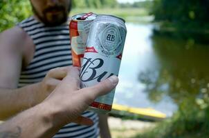 Young man raise Budweiser Bud beer can with male friend on blurred river with kayak and trees photo