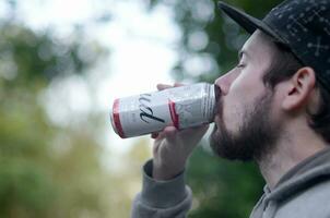 Young bearded man drinks Budweiser Bud beer in aluminium can on green trees blurred background photo