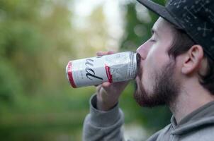 Young bearded man drinks Budweiser Bud beer in aluminium can on green trees blurred background photo