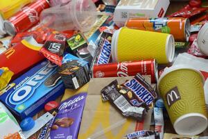 Big pile of many disposable colored trash from edible products of famous food and beverage manufacturers. Soda cans and chocolate bar wrappings in stack photo