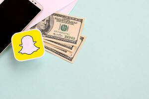 Snapchat paper logo lies with envelope full of dollar bills and smartphone photo