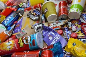 Big pile of many disposable colored trash from edible products of famous food and beverage manufacturers. Soda cans and chocolate bar wrappings in stack photo