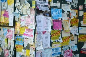 KHARKOV, UKRAINE - OCTOBER 2, 2019 Grunge Message Board with many advertisement. People spreading advertising and information on white paper than post it on metal fence photo