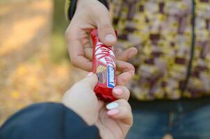 Jarkov, Ucrania - octubre 21, 2019 masculino mano pasa el niña un equipo Kat chocolate bar en un otoño parque. el manifestación de amabilidad, tratando con dulces foto