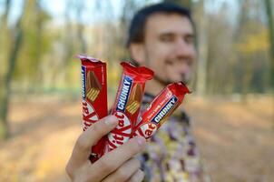 Jarkov, Ucrania - octubre 21, 2019 un joven caucásico barbado hombre muestra Tres equipo Kat chocolate barras en rojo envase en otoño parque. equipo Kat chocolate bienes fabricado por hacerse un nido foto