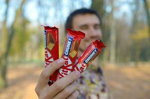 KHARKOV, UKRAINE - OCTOBER 21, 2019 A young caucasian bearded man shows three kit kat chocolate bars in red wrapping in autumn park. Kit Kat chocolate goods manufactured by Nestle photo