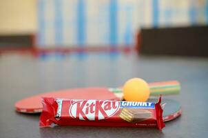 KHARKOV, UKRAINE - OCTOBER 17, 2019 Kit kat chocolate bar in red wrapping next to ping pong rackets and balls on table in a outdoor sport yard photo