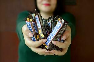 KHARKOV, UKRAINE - OCTOBER 8, 2019 A young caucasian brunette girl shows many Snickers chocolate bars in dark room. Snickers chocolate manufactured by Mars photo