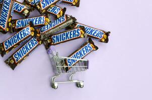 Many Snickers chocolate bars stacked close up with shopping cart on light violet background. Snickers bars are produced by Mars Incorporated photo