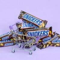 Many Snickers chocolate bars stacked close up with shopping cart on light violet background. Snickers bars are produced by Mars Incorporated photo