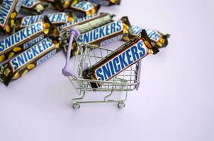 Many Snickers chocolate bars stacked close up with shopping cart on light violet background. Snickers bars are produced by Mars Incorporated photo