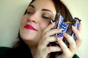 KHARKOV, UKRAINE - OCTOBER 8, 2019 A young smiling caucasian brunette girl shows many Snickers chocolate bars in dark room. Snickers chocolate manufactured by Mars photo