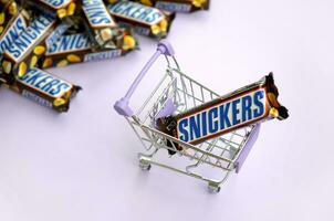 Many Snickers chocolate bars stacked close up with shopping cart on light violet background. Snickers bars are produced by Mars Incorporated photo