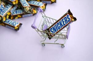 Many Snickers chocolate bars stacked close up with shopping cart on light violet background. Snickers bars are produced by Mars Incorporated photo