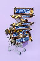 Many Snickers chocolate bars stacked close up with shopping cart on light violet background. Snickers bars are produced by Mars Incorporated photo