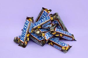 Many Snickers chocolate bars lies on pastel lilac paper. Snickers bars are produced by Mars Incorporated. Snickers was created by Franklin Clarence Mars in 1930 photo