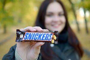 KHARKOV, UKRAINE - OCTOBER 8, 2019 A young caucasian brunette girl shows Snickers chocolate bar in autumn park. Snickers chocolate manufactured by Mars photo