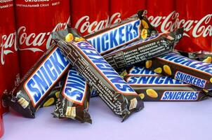 Snickers chocolate bars in brown wrapping lies on bright violet background with Coca Cola tin cans close up. Famous drink and chocolate product photo