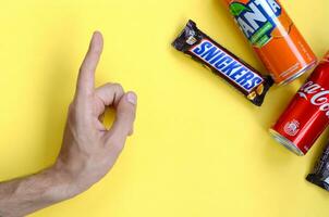 Male hand points on Snickers chocolate bar in brown wrapping, red Coca Cola and orange Fanta tin can on yellow pastel background. Famous drink and chocolate product photo