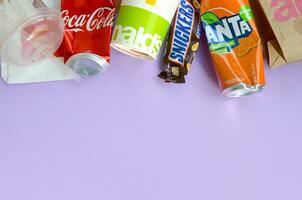 Snickers chocolate bar wrapping lies with empty red Coca Cola and orange Fanta tin cans next to mcdonalds paper bags and cups on lilac pastel background. Famous drink and chocolate product trash photo