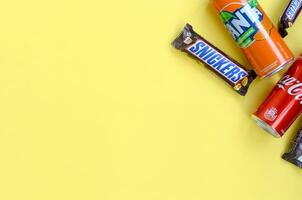 Snickers chocolate bar in brown wrapping lies with red Coca Cola and orange Fanta tin can on yellow pastel background. Famous drink and chocolate product photo