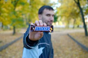 KHARKOV, UKRAINE - OCTOBER 8, 2019 A young caucasian bearded man shows Snickers chocolate bar in brown wrapping in autumn park. Snickers chocolate manufactured by Mars photo