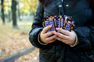 KHARKOV, UKRAINE - OCTOBER 8, 2019 A young caucasian woman shows many Snickers chocolate bars in brown wrapping in autumn park. Snickers chocolate manufactured by Mars photo