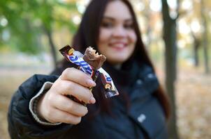 KHARKOV, UKRAINE - OCTOBER 8, 2019 A young caucasian brunette girl shows Snickers chocolate bar in autumn park. Snickers chocolate manufactured by Mars photo