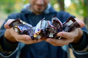 KHARKOV, UKRAINE - OCTOBER 8, 2019 A young caucasian bearded man shows many Snickers chocolate bars in brown wrapping in autumn park. Snickers chocolate manufactured by Mars photo