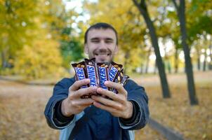 KHARKOV, UKRAINE - OCTOBER 8, 2019 A young caucasian bearded man shows many Snickers chocolate bars in brown wrapping in autumn park. Snickers chocolate manufactured by Mars photo
