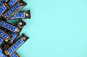 Many Snickers chocolate bars lies on pastel blue paper. Snickers bars are produced by Mars Incorporated. Snickers was created by Franklin Clarence Mars in 1930 photo