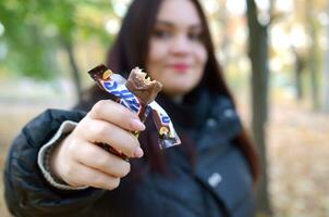 KHARKOV, UKRAINE - OCTOBER 8, 2019 A young caucasian brunette girl shows Snickers chocolate bar in autumn park. Snickers chocolate manufactured by Mars photo