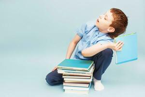 schoolboy sitting with pile of school books and throwing a textbook isolated on a blue background photo