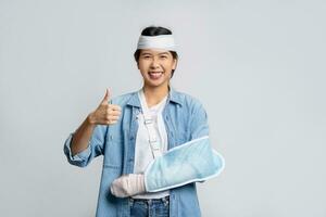 Smiling asian woman with broken head and wearing soft cast due to broken arm while showing thumbs up isolated on white background. Medical expenses concept. photo