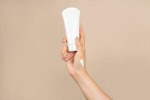 Female hand with cream to touch cosmetic tube on beige background. Concept of hand lotion cream or winter facial care. photo