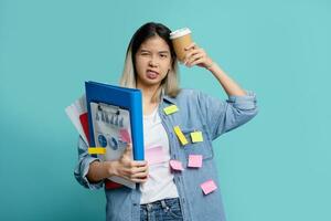 Bored Asian female office worker with a lot of work with holding document file  with cup of coffee and note on face standing on blue background in studio. photo