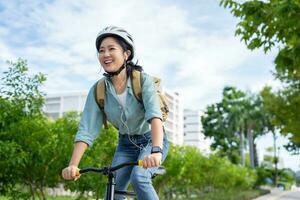 contento asiático mujer vistiendo un casco y escuchando a su favorito música mientras montando un bicicleta mediante un ciudad parque. foto